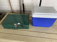 Closed green Coleman two-burner camping stove and blue Thermos cooler box side by side