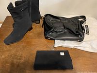 Pair of black Gore Tex boots, black leather purse on Soprano branded white cloth, and small black clutch purse on wooden table.
