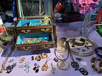 Open wooden jewelry box with blue velvet lining showing multiple compartments filled with various costume jewelry items. Jewelry includes necklaces, brooches, earrings, and pendants of different styles and colors.