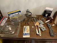 Full lot of kitchen items including glass casseroles, vintage cookie cutters, wooden utensils, egg beater, French press, travel mug and tape dispenser arranged on a table.