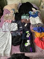 Overview of the full clothing lot laid on bed showing various tops, bathing suit tops, jeans, skirts, tights, and packaged undergarments