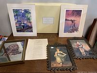 Six framed pictures on a table including two Robert Harms prints in white frames, one new Pottery Barn frame, two ornate vintage Italian framed pictures, and one mirrored owl picture.