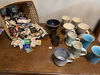 Wide view showing all items: basket of matchboxes tipped over spilling matches, a dark bowl, and eleven assorted pottery mugs in various glazes and colors.
