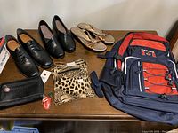 Full view of the lot items on table showing two pairs of shoes size 8, one pair of Amalie sandals size 8.5, a Canada backpack, a black leather wallet, and a leopard print purse