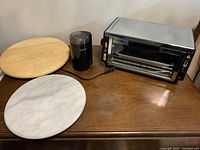 Photo showing Proctor Silex toaster oven, black Cuisinart coffee grinder, wooden and marble Lazy Susans on a wooden table