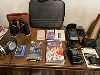 Full view of lot items on table including cameras, VHS tapes, Walkman, headphones, binoculars, cases, instruction manuals.