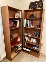 Two solid wood bookshelves placed side by side in a room, each with several books and some decorative items on the shelves. Clean lines and medium brown wood finish.
