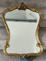 Front view of decorative wall mirror showing ornate gold-tone frame with scroll and floral detail and clear mirror glass.