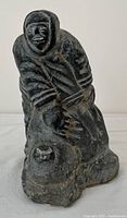 Photo of dark stone Inuit carving showing front view of a figure wearing hooded parka clothing with carved facial features and base.