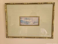 Frontal photo of the framed Japanese scene artwork, showing the delicate six narrow panels arranged horizontally with a subtle traditional painting of figures and water, surrounded by a bamboo-style frame.