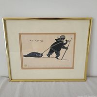 Framed print showing an Inuit figure pulling a seal, signed by Henry Napartok with matting and gold-colored metal frame.