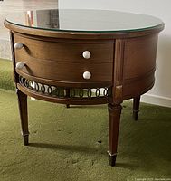 Round wooden corner table with clear glass top and two drawers, shown from an angle highlighting the drawers and decorative metal rail below.