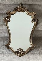 Front view of a decorative hanging mirror showing ornate gold-tone frame with floral and scroll details, mirror surface clear and reflective.