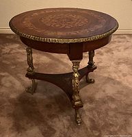 Angled front view of round table showing inlaid top, gilt metal trim, and carved tripod legs.