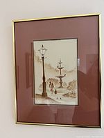 Framed watercolor painting showing Parisian park scene with lamp post, fountain, and figures walking; reddish-brown matte and gold-tone frame