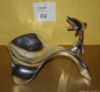 Single polished metal goose sculpture on yellow background, labelled with auction tag