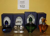 Four crystal glass perfume containers displayed in front of their original blue boxes with satin white interiors. The containers vary in color: dark blue, clear, green, and amber orange, all approximately 3 1/2 inches tall, with faceted, geometric designs and matching stoppers.