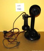 Front view of black antique candlestick telephone showing the vertical stand with mouthpiece, round base, connected handset lying beside it, original wiring with plug and some fraying