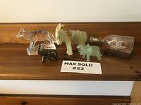 Lot of five miniature animal figurines including stone onyx donkey and elephant, brass elephant, glass fox, and a ship in a bottle on wooden base, all arranged on a wooden shelf.
