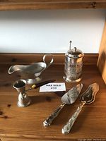 Overview of the lot showing all five items on a wooden surface, including gravy boat, candle snuffer, jam jar cover, and two cake servers.