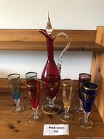 Full view of tall red vintage glass decanter with pointed stopper and applied twisted handle, surrounded by six colorful wine glasses in blue, green, red, amber, purple, and dark blue colors with gold rims and detailing.