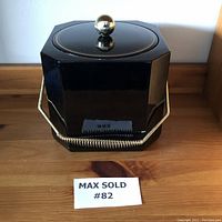 Front view of a black lacquer hexagonal ice bucket with gold-tone round knob lid and metal handle, showing slight wear at handle attachment points.