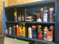 Blue metal cabinet shelf with an assortment of automotive sprays and liquids including Zep 45 Cleaner, STP, Fast Orange hand cleaner, brake parts cleaner, clear coat, and 3 in 1 oil.