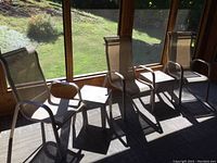 Three metal framed patio chairs and two resin side tables arranged on a patio with wooden walls and large windows.