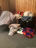 Variety of folded and stacked bed coverings, quilts, and blankets on a chair and floor in wood-paneled room.