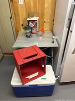 View of folding table with items including Coleman lantern, flashlight, two red milk crates, white bucket, and Coleman cooler underneath.