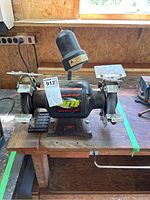 Front view showing the main body of the Trademaster TMT bench grinder sitting on a wooden workbench, with a lamp mounted on top and protective guards over grinding and wire brush wheels.