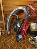 Hoover WindTunnel canister vacuum with power head and hose, red mesh bag attached to the handle, sitting on brown carpet against wood panel wall.