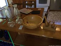 Wide shot showing pair of glass oil lamps, wooden bowl, and carved birds on a wooden table.