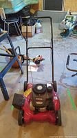 Full view of the red push lawn mower with black handle on indoor concrete floor showing front and top engine area.
