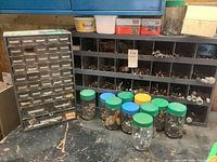 Overview of two metal storage caddies with numerous small drawers filled with assorted nuts, bolts, and screws along with plastic jars containing more hardware items on a workbench.