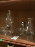 Both oil lamps shown on wooden shelf: left with finger hold, right with rounded bottom and chimneys.