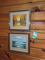 Two framed oil paintings hanging on wood wall, cityscape above ocean scene, both 8x10 inches, signed