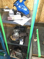 Blue electric chainsaw sharpener with a vice clamp, accessories, and a manual on gray shelving.
