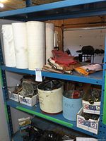 Shelf with six white rolls of shop towels, red and brown work gloves, and various tins and small boxes of hardware items including door knobs and clamps.