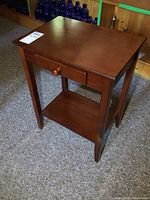 Full view of the modern wood side table showing the smooth top surface, small front drawer with round wooden knob, and lower shelf supported by four tapered legs.
