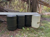 Four plastic/resin water barrels under metal sheet, black and white colors, stored outdoors by trees on grass.