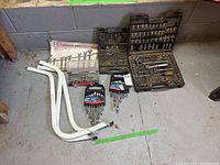 Full lot showing socket set, bit sets, combination wrench sets, and three metal table legs on concrete floor against wall