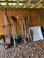Full view of lot including snow scoop, shovels, garden tools leaning against wood wall on gravel floor.