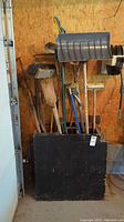 Full view of the black painted wooden storage container holding multiple yard tools including snow shovel, brooms, axe, and squeegee