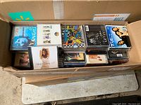 Overhead view of an open cardboard box filled with approximately 120 compact discs in jewel cases, displaying diverse cover art.