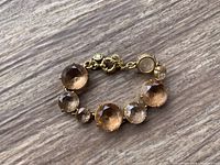Close-up photo of a gold-tone bracelet featuring round faceted brown and clear stones set alternately with small chain links and a round spring clasp, placed on a wooden surface.