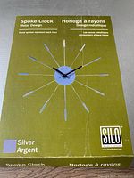 Photo of the SILO Spoke Clock box showing the product name, color (silver) and design details on an olive green background.