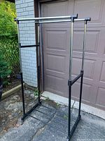 Metal clothing rack with adjustable double hanging rods and lower shelf shown outdoors against a garage door and brick wall.