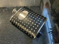 Photo showing metal flask covered in black leather with silver dots and a silver engraving plaque on front, placed on black granite countertop.