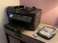 Black HP colour multifunction printer placed on a wooden desk next to cables, CDs, and a small cartridge or accessory.